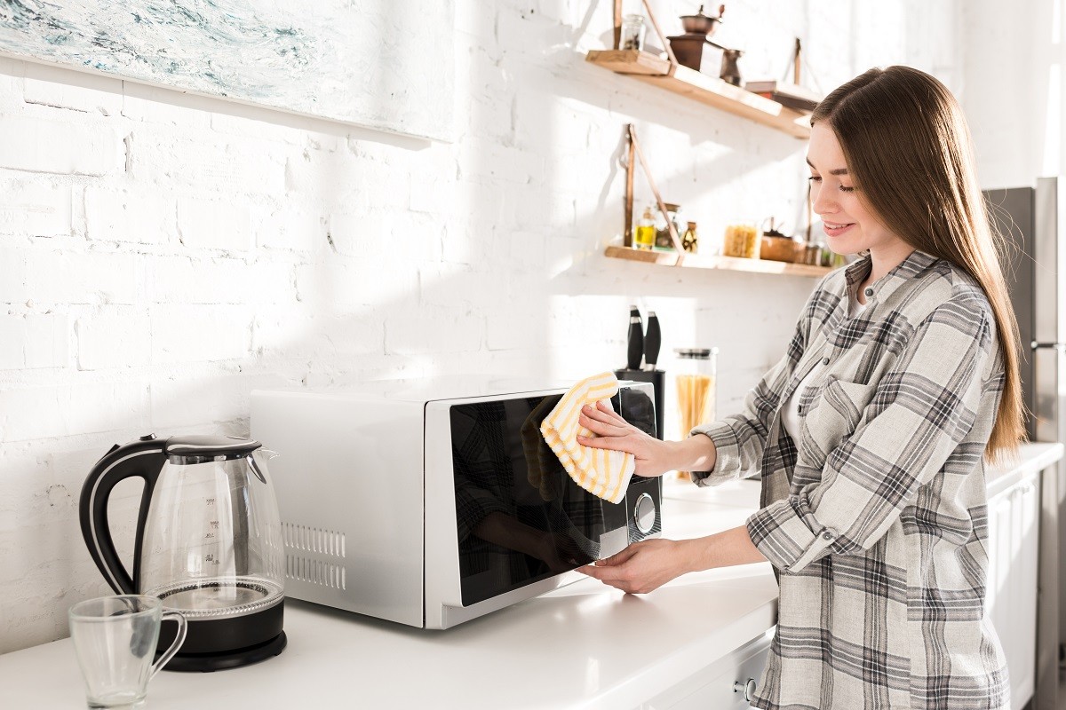 smiling and attractive woman cleaning microwave with rag in kitchen