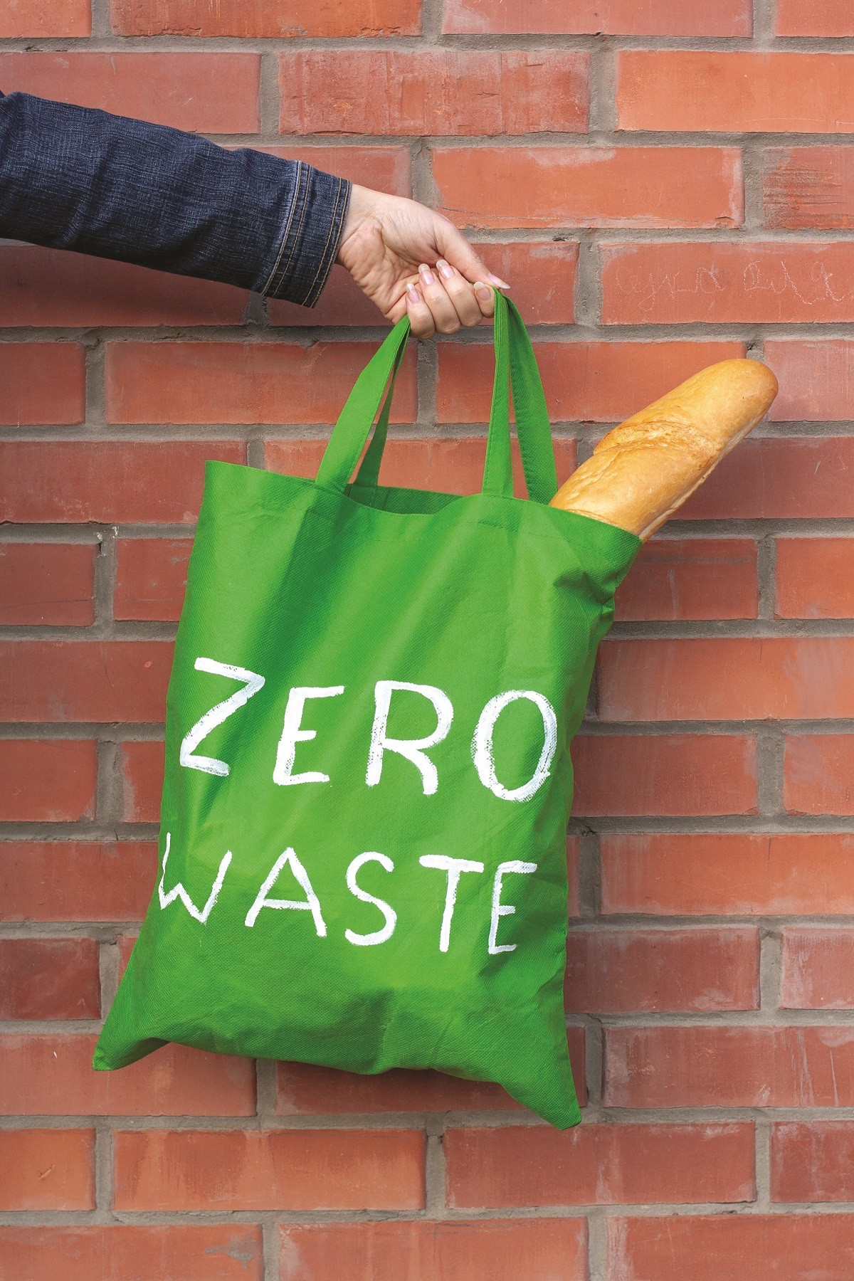 Green eco bag with a white inscription zero waste which the hand holds on a background of a brick wall.