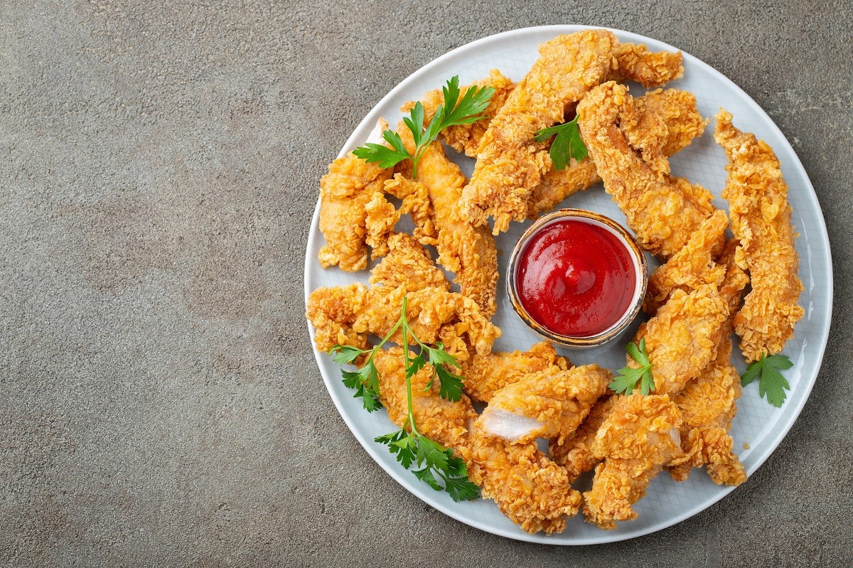 Breaded chicken strips with tomato ketchup on a white plate. Fast food on dark brown background. Top view with copy space