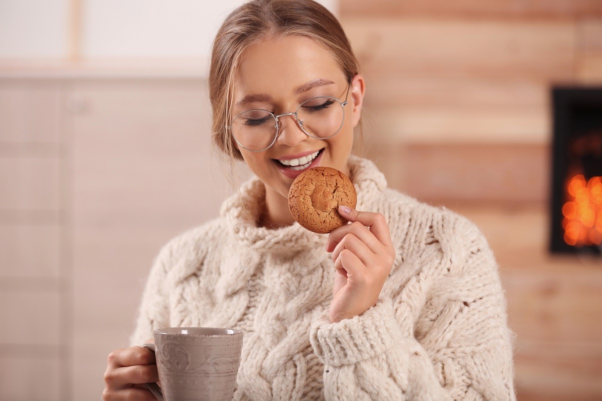 Beautiful young woman with cup of hot drink and cookie at home. Winter atmosphere