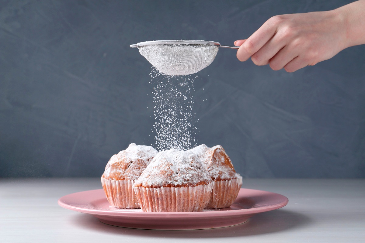 Woman,With,Sieve,Sprinkling,Powdered,Sugar,Onto,Muffins,At,White